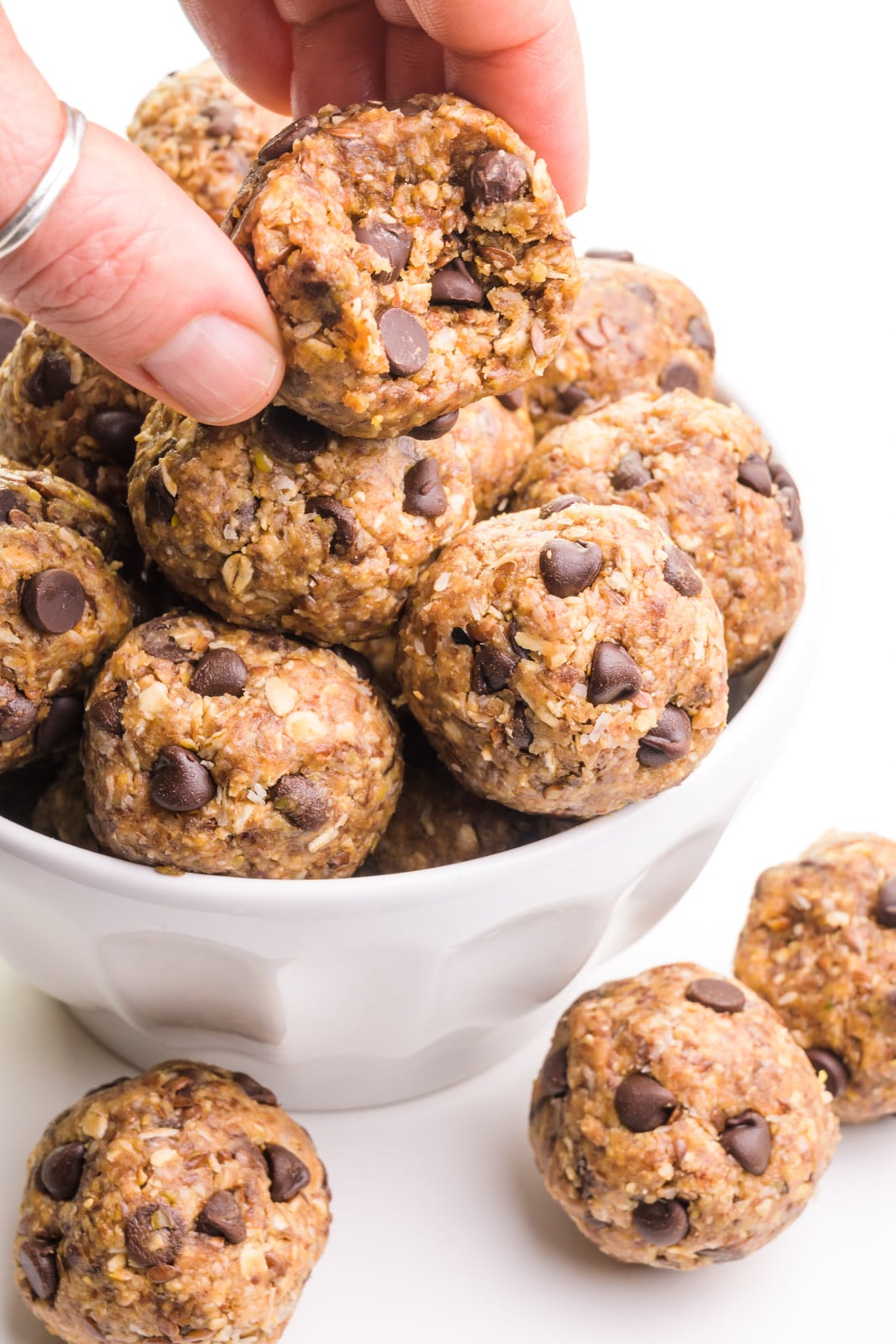 A hand holds an energy ball with a bite taken out, hovering over a bowl with more of the energy balls. There are three of the treats on the table beside the bowl.