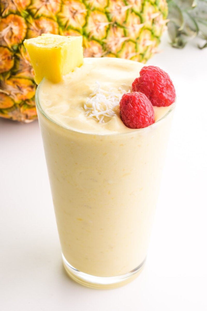 A glass full of Pina colada smoothie has raspberries, coconut flakes, and a pineapple chunk on top. It sits in front of a pineapple.
