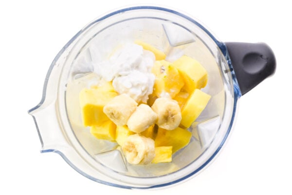 Looking down on a blender full of pineapple chunks, banana chunks, coconut milk, and more.
