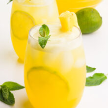Two glasses sit one in front of the other. Both hold pineapple drinks with fresh limes, mint sprigs, and pineapple chunks. There are more mint herbs, a lime, and a pitcher with more of the beverage in the background.