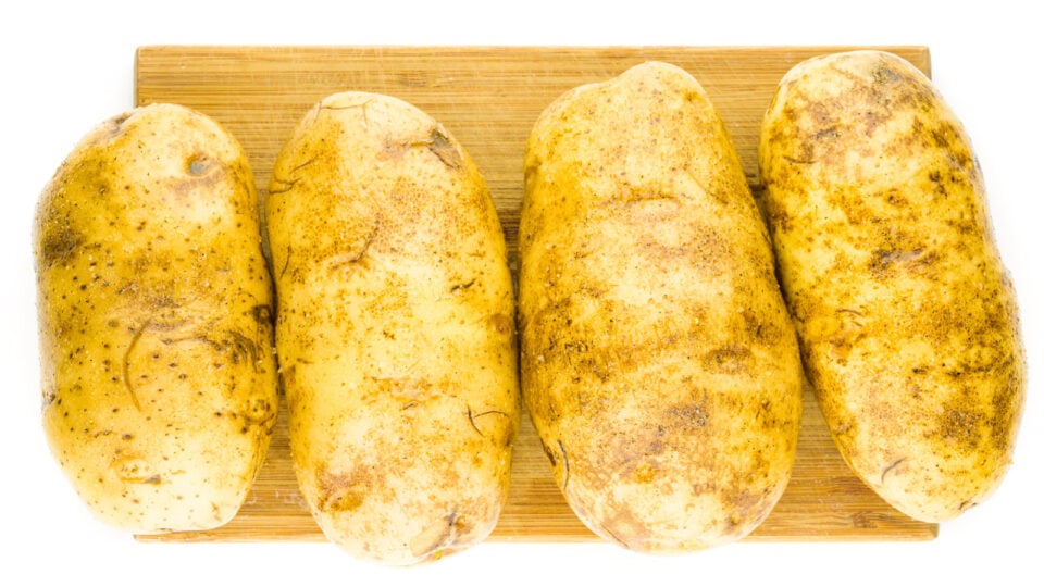 Four potatoes have been picked with a fork, covered with oil, and seasoned with salt and pepper. They are sitting on a cutting board.