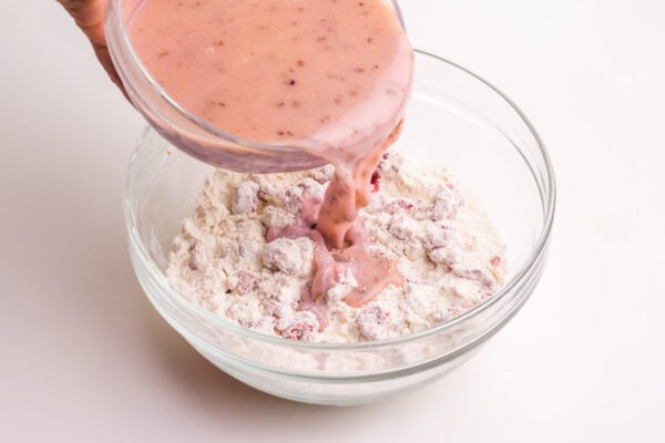 A hand holds a bowl pouring a pink milk mixture into a bowl with flour.