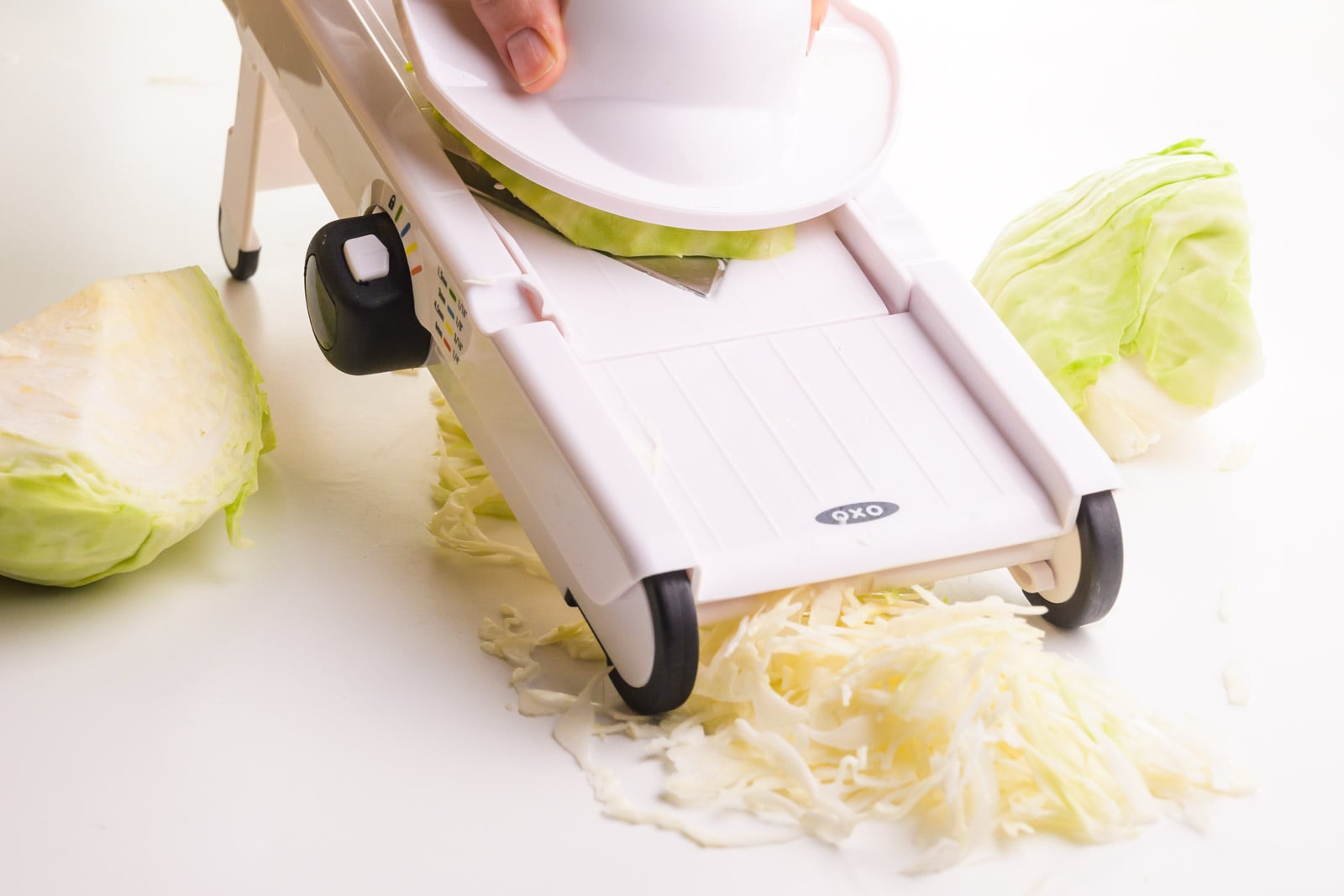 A hand holds a tool shredding green cabbage with a mandoline. There are large pieces of green cabbage beside it.