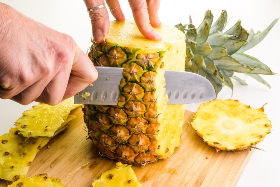The top and bottom of a fresh pineapple have been chopped off. A hand holds a knife, cutting down the outer edge of the pineapple to remove the outer layer.