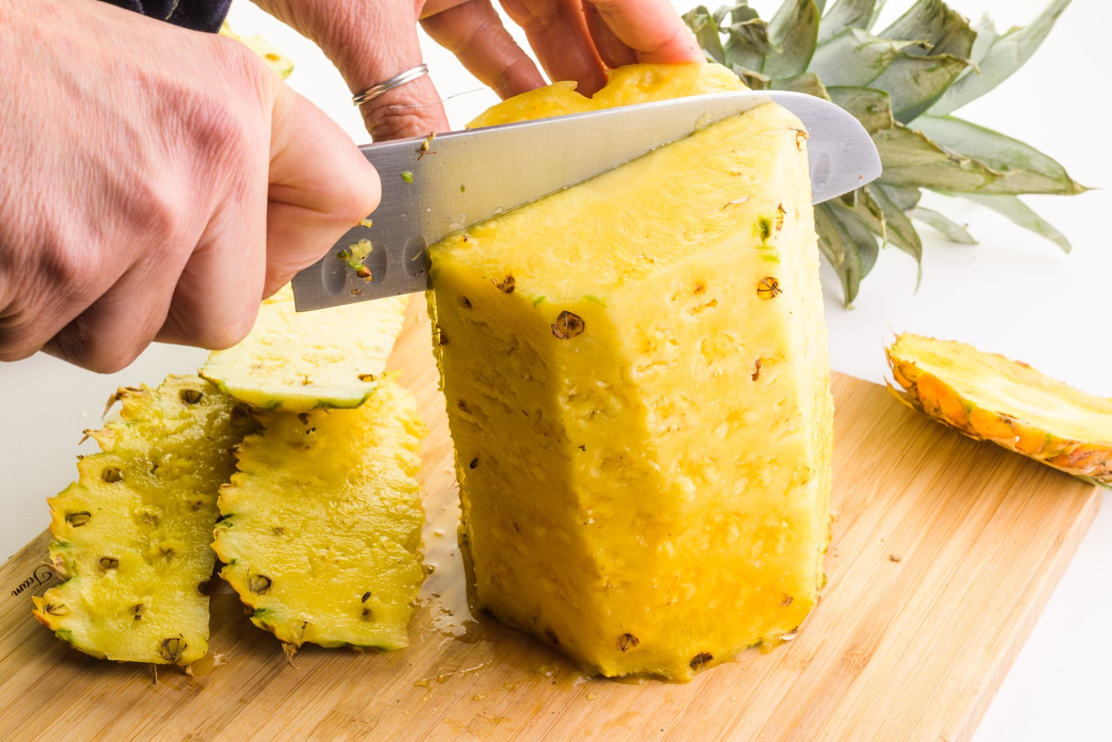 A hand holds a knife cutting down on a skinned pineapple. There are pieces of the pineapple skins sitting around it on a cutting board.