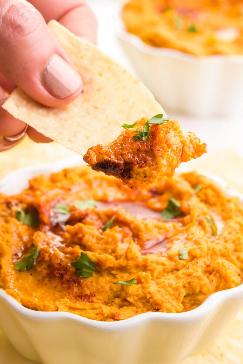 A hand holds a tortilla chip with sweet potato dip on it. It hovers over a bowl with more of the dip and there's another bowl in the background.