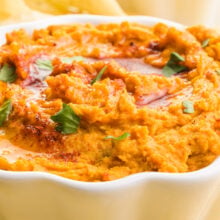 A close-up shot of sweet potato hummus in a bowl. It's topped with chopped cilantro, olive oil, and paprika. There's another bowl and tortilla chips in the background.