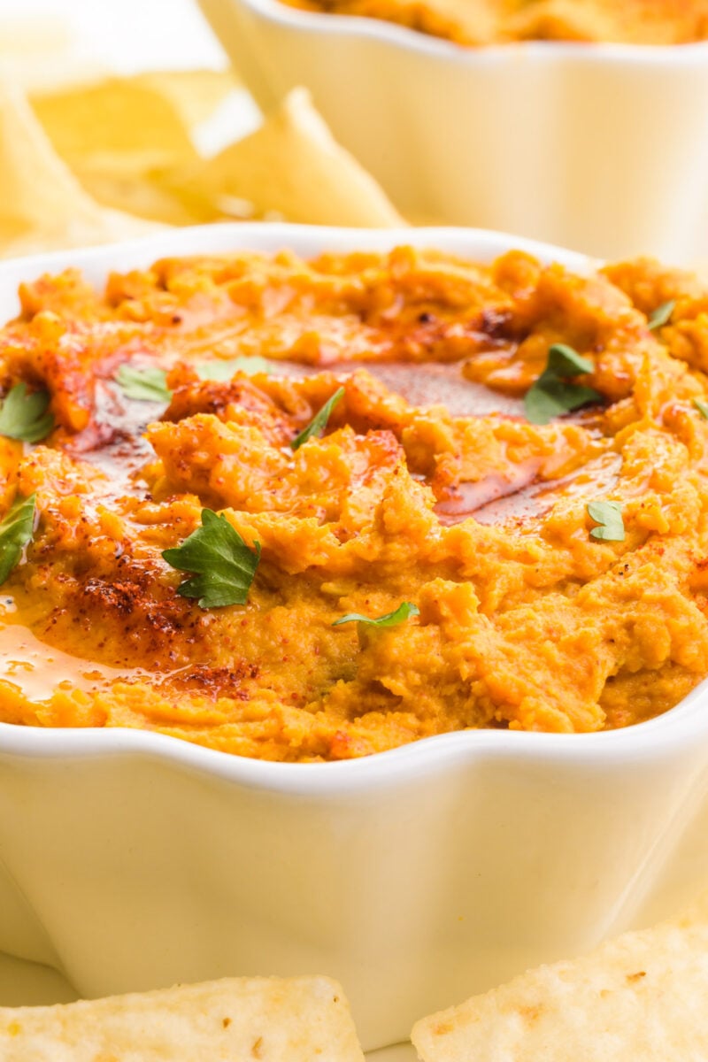 A close-up shot of sweet potato hummus in a bowl. It's topped with chopped cilantro, olive oil, and paprika. There's another bowl and tortilla chips in the background.