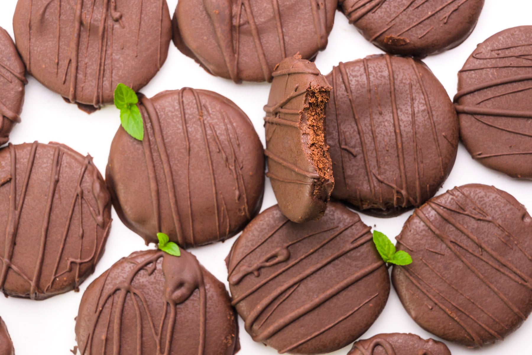 Looking down on several chocolate cookies sitting next to it. One is tilted up and has a bite taken out of it. There are mint sprigs around the cookies.