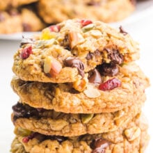 A stack of trail mix cookies shows the top one with a bite taken out. There are more cookies in the background.