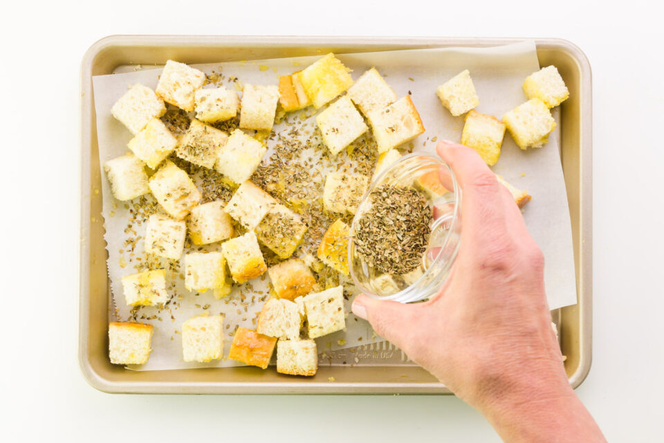 A hand holds a small bowl of seasoning, pouring it over bread cubes in a pan.