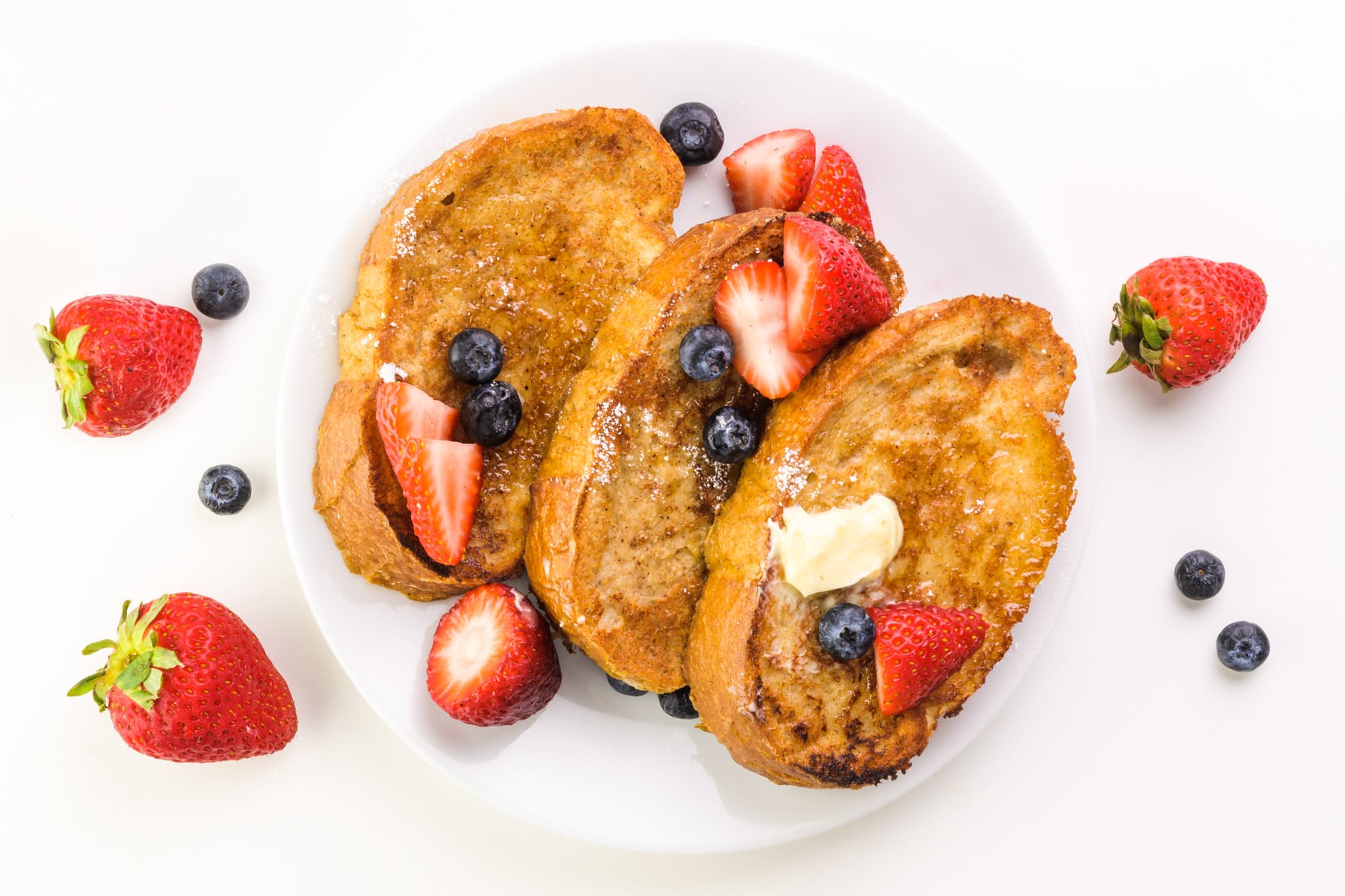 Looking down on a plate with slices of French toast topped with blueberries, strawberries ,and butter.