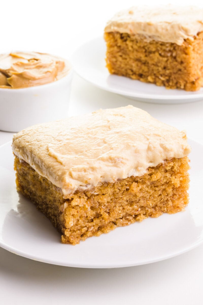 A slice of vegan peanut butter cake sits on a plate, there's a another slice and a bowl of peanut butter in the background.