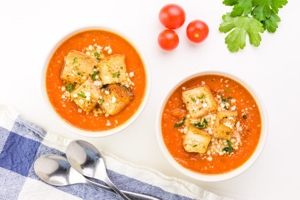 Looking down on two bowls of soup, both with croutons on top. There are fresh tomatoes and herbs nearby. There are two spoons on a napkin by the bowls.