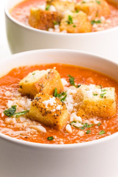 A bowl of vegan tomato soup has croutons and herbs on top. There is another bowl in the background.