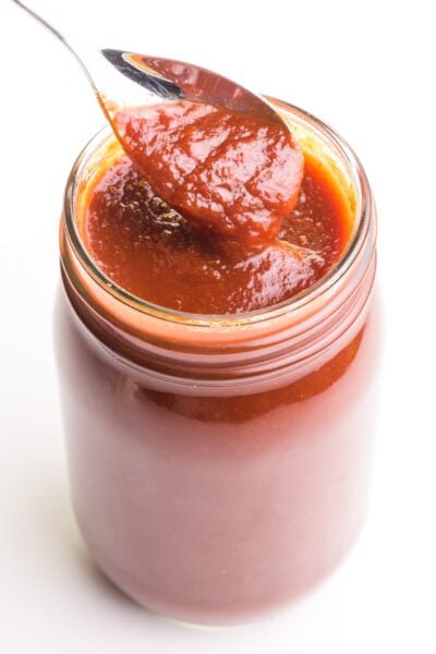 A spoon holds vegan bbq sauce over a glass jar full of the sauce.