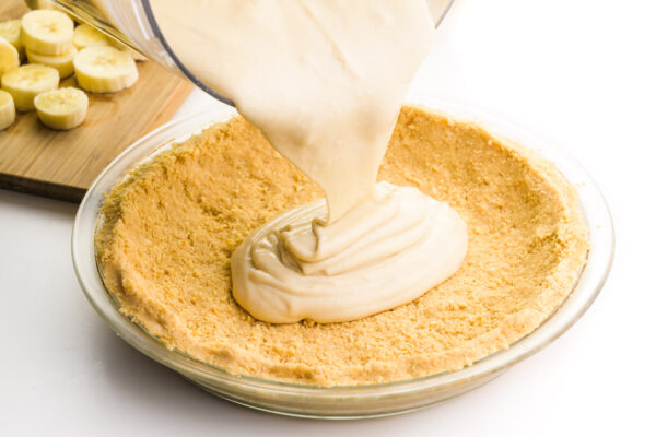 Vanilla pudding is being poured into a golden cookie crust. There is a cutting board with sliced bananas in the background.
