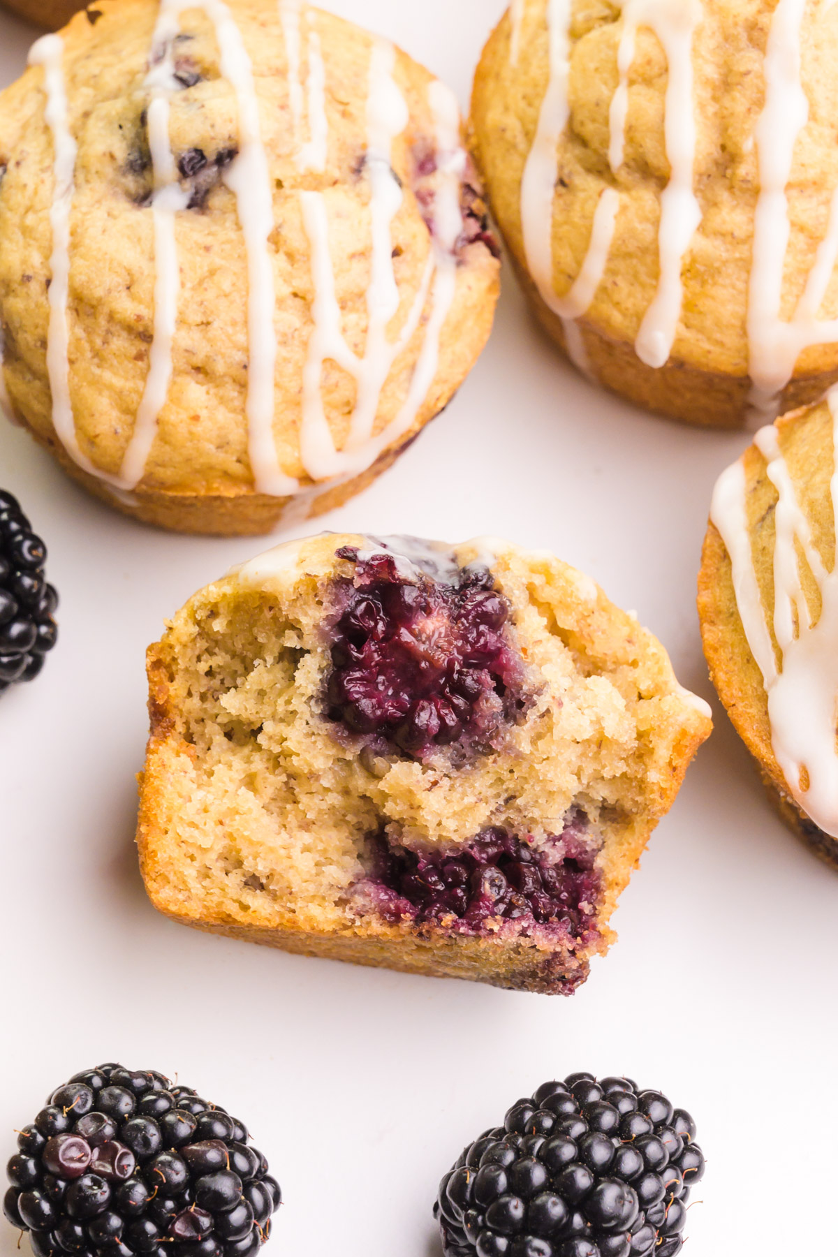 Looking down on a muffin with a bite taken out. It shows big blackberries inside. There are other muffins around it along with fresh blackberries.