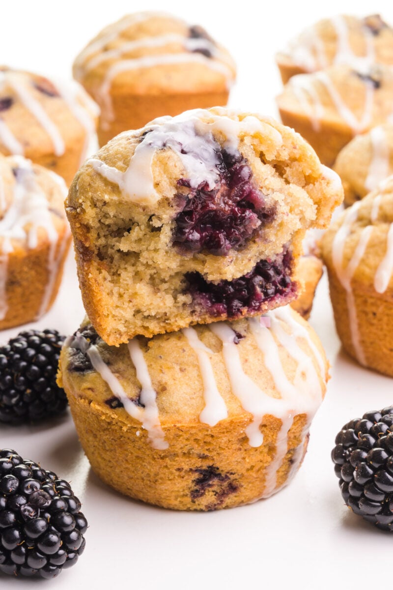 A blackberry muffin with a bite out of it sits on top of another muffin. There are fresh blackberries and more muffins around the stack.
