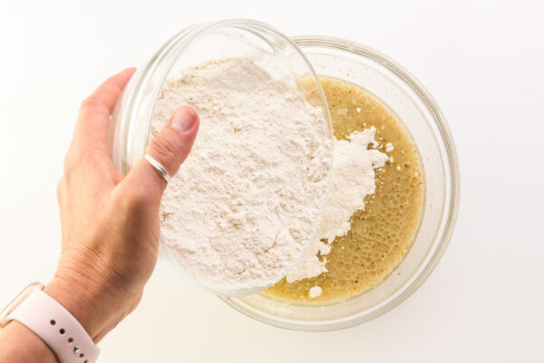 A hand holds a bowl pouring flour into another bowl with wet ingredients.