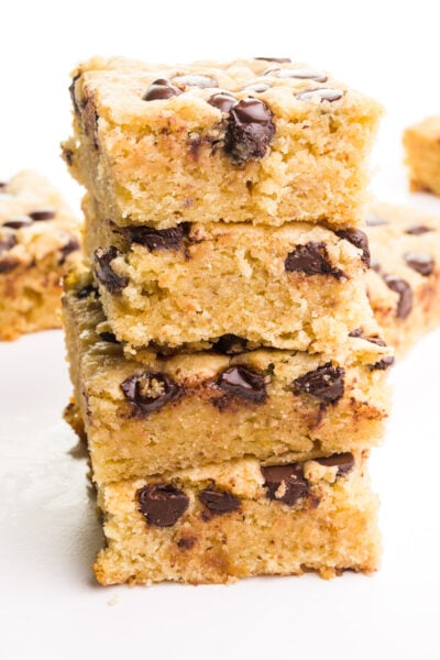 A stack of vegan blondies shows chocolate chips on top and on the sides. There are more slices in the background.