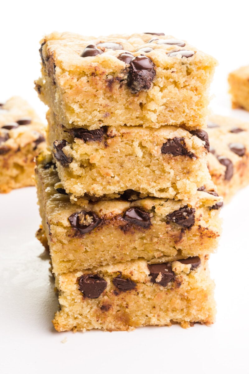 A stack of vegan blondies shows chocolate chips on top and on the sides. There are more slices in the background.