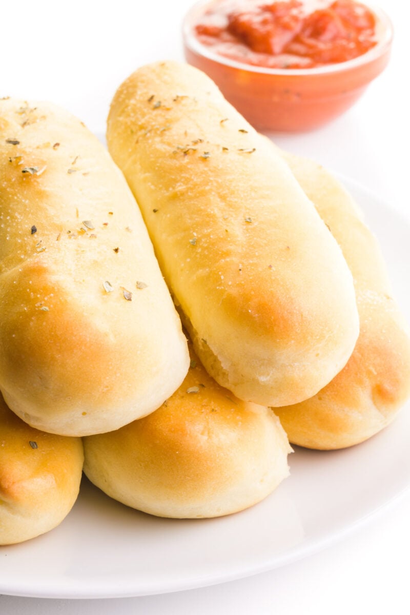 Vegan breadsticks are piled high on a plate sitting in front of a bowl of marinara sauce.