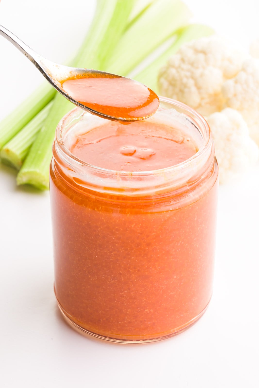 A spoon full of vegan buffalo sauce hovers over the rest of the jar. There are celery sticks and cauliflower florets in the background.