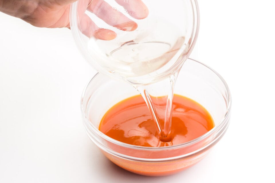 A. hand holds a bowl of melted coconut oil, pouring it into another bowl with red pepper sauce.