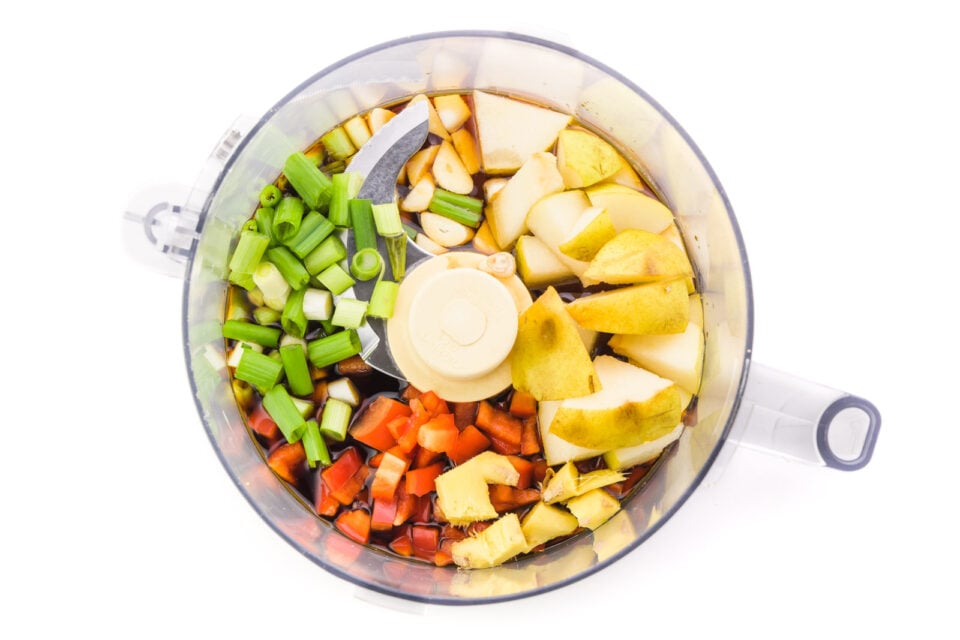 Ingredients, like green onions, red bell peppers, and fresh ginger, are in the bottom of a food processor bowl.