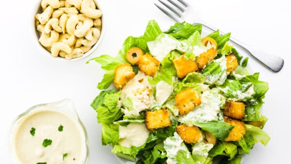 Looking down on a vegan caesar salad with a fork, dressing, and a bowl of cashews next to it.