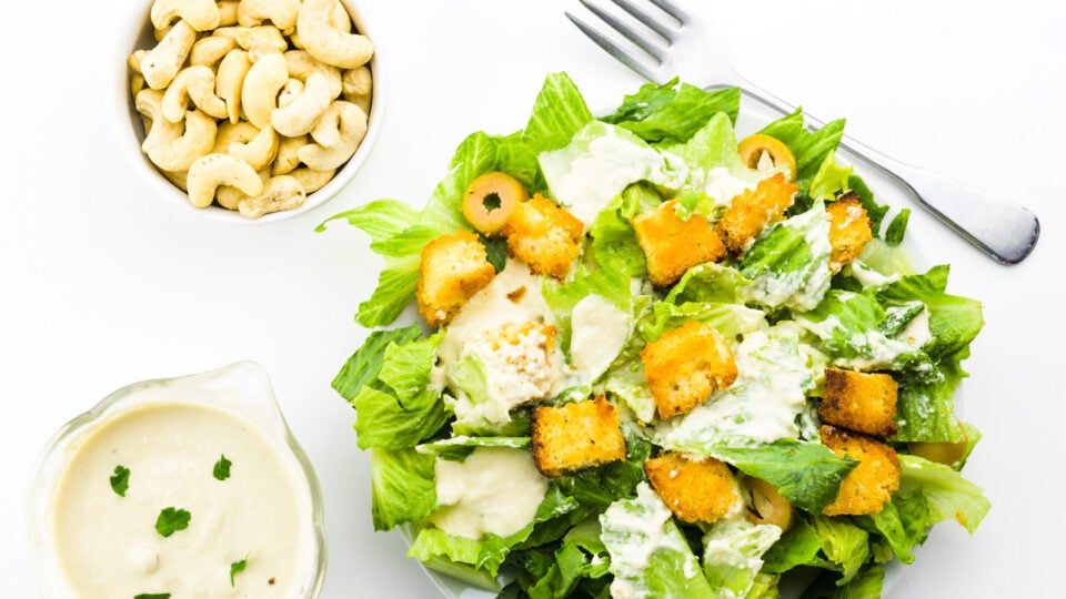 Looking down on a vegan caesar salad with a fork, dressing, and a bowl of cashews next to it.