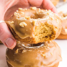 A hand holds a caramel donut hovering over another donut. There are more in the background, too.