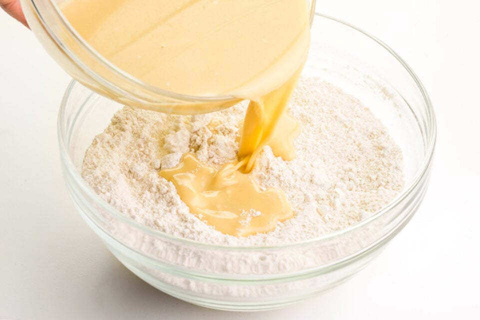 A bowl holds a liquid mixture and is pouring it into another bowl full of flour.