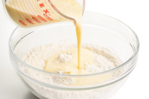 A liquid milky mixture is being poured into a glass bowl full of flour.