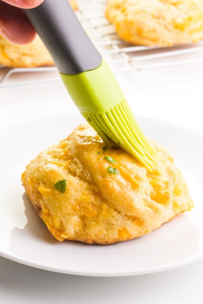 A hand holds a pastry brush, spreading topping over a biscuit. There are more biscuits in the background.