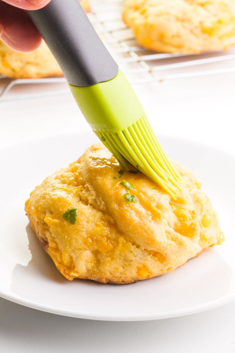 A hand holds a pastry brush, spreading topping over a biscuit. There are more biscuits in the background.