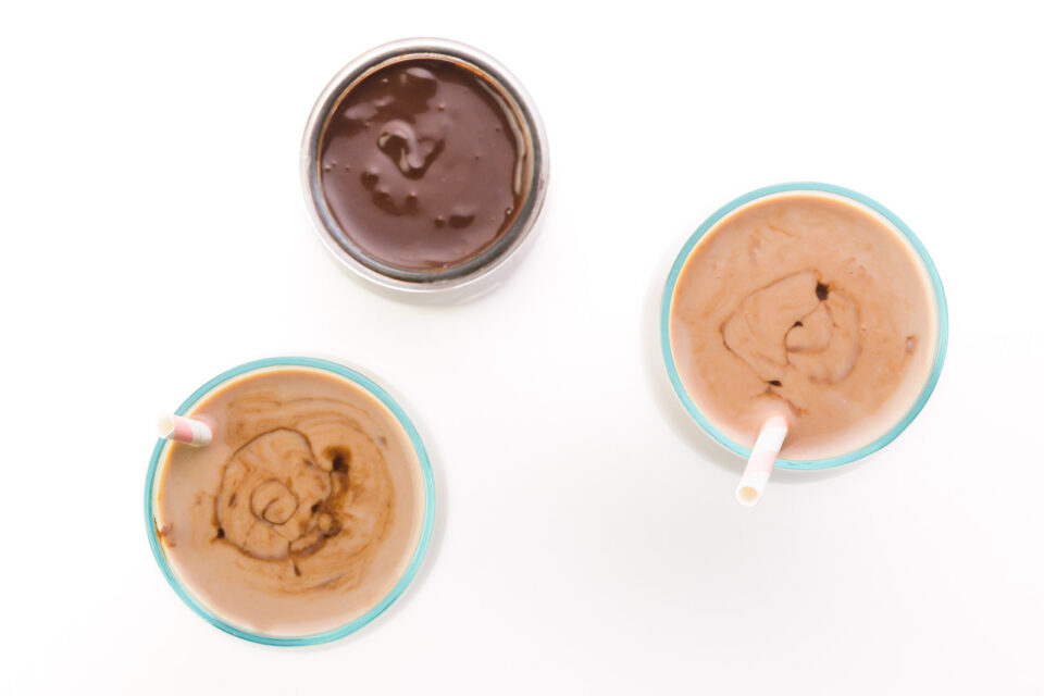 Looking down on two glasses of chocolate milk, both with straw in the glass. There is a glass jar of chocolate syrup between them.