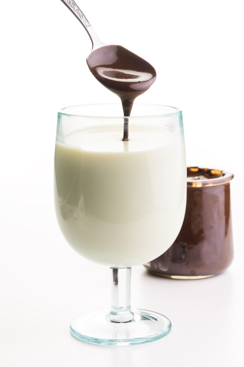 A spoon is pouring chocolate syrup into a glass of soy milk. The jar of chocolate syrup in the background.