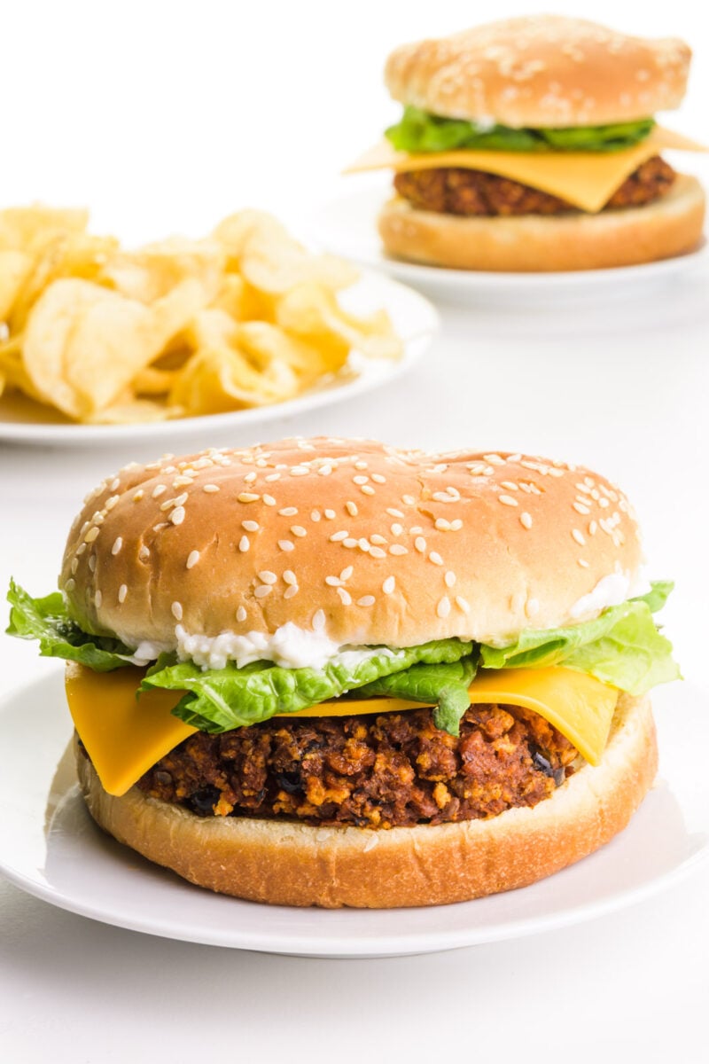 A vegan chorizo burger sits on a plate with a plate of potato chips in the background. There's another burger in the background too.