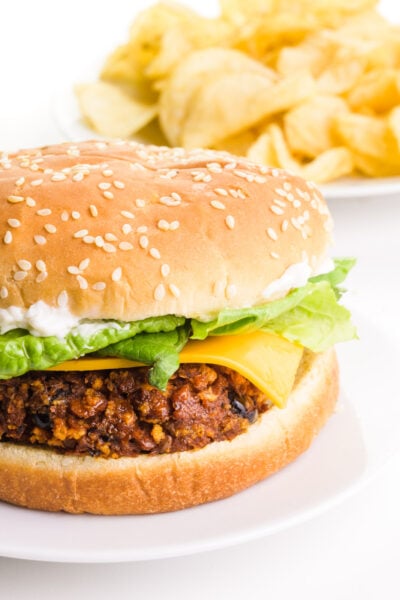 A vegan chorizo burger sits on a plate with a plate of potato chips in the background.