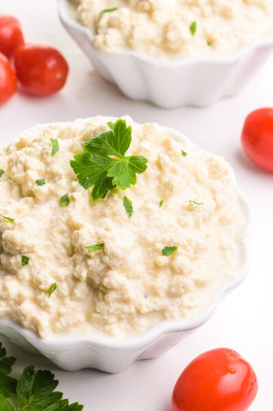 A bowl of vegan cottage cheese has green herbs on top. There's another bowl in the background, along with cherry tomatoes and more fresh herbs.