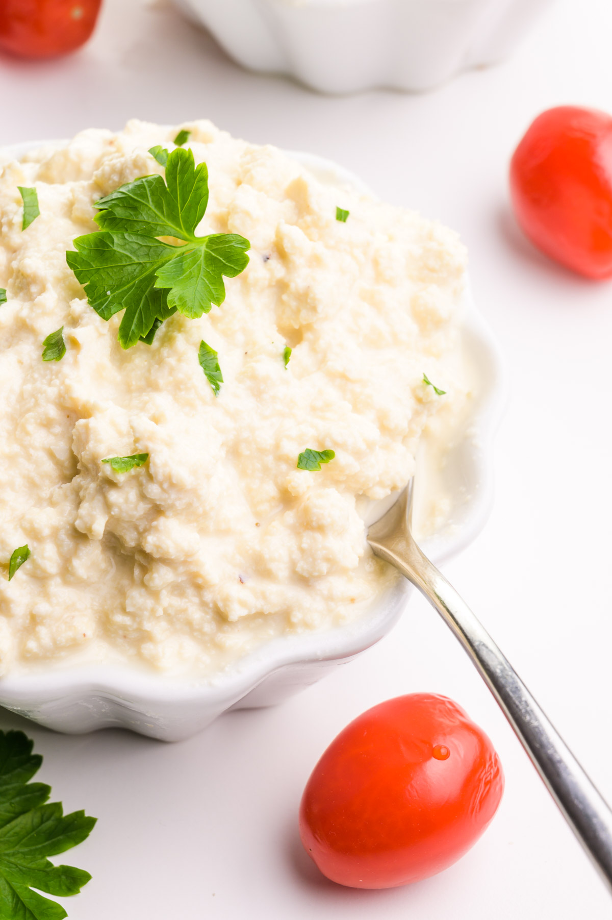 A bowl of cottage cheese has a spoon in it and green herbs on top of it. There is another bowl in the background and cherry tomatoes and more green herbs around it.