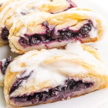 A vegan danish sits on a plate. It has blueberry filling and vanilla glaze on top.