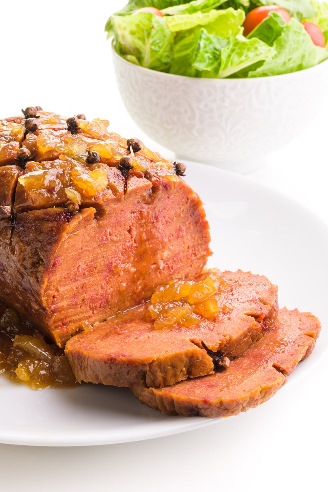 A vegan ham loaf has two slices cut out on a plate. There is pineapple sauce on top of the slices ad the loaf. There's a salad in the background.