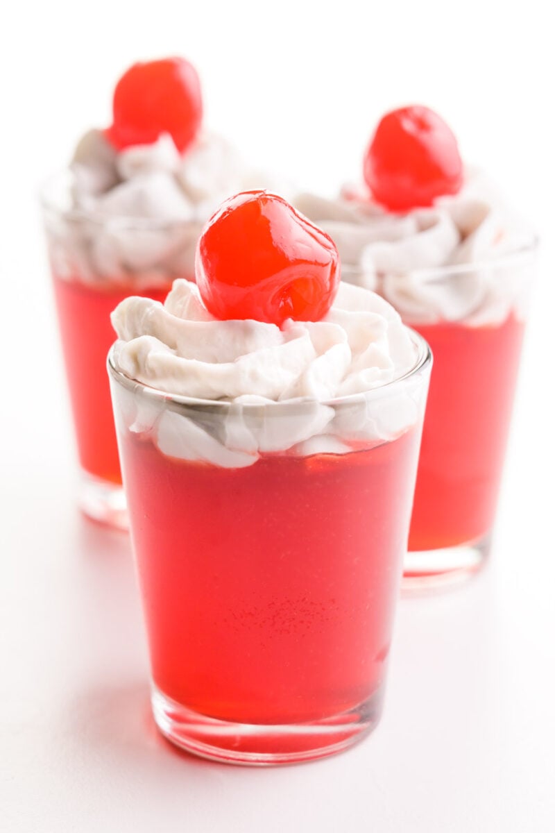 Three shot glasses have cherry vegan jello shots that are topped with whipped cream and maraschino cherries.