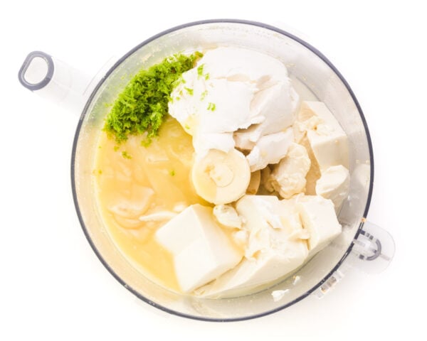 Looking down into a food processor bowl with ingredients like vegan cream cheese, silken tofu, sweetened condensed milk, and key lime zest.