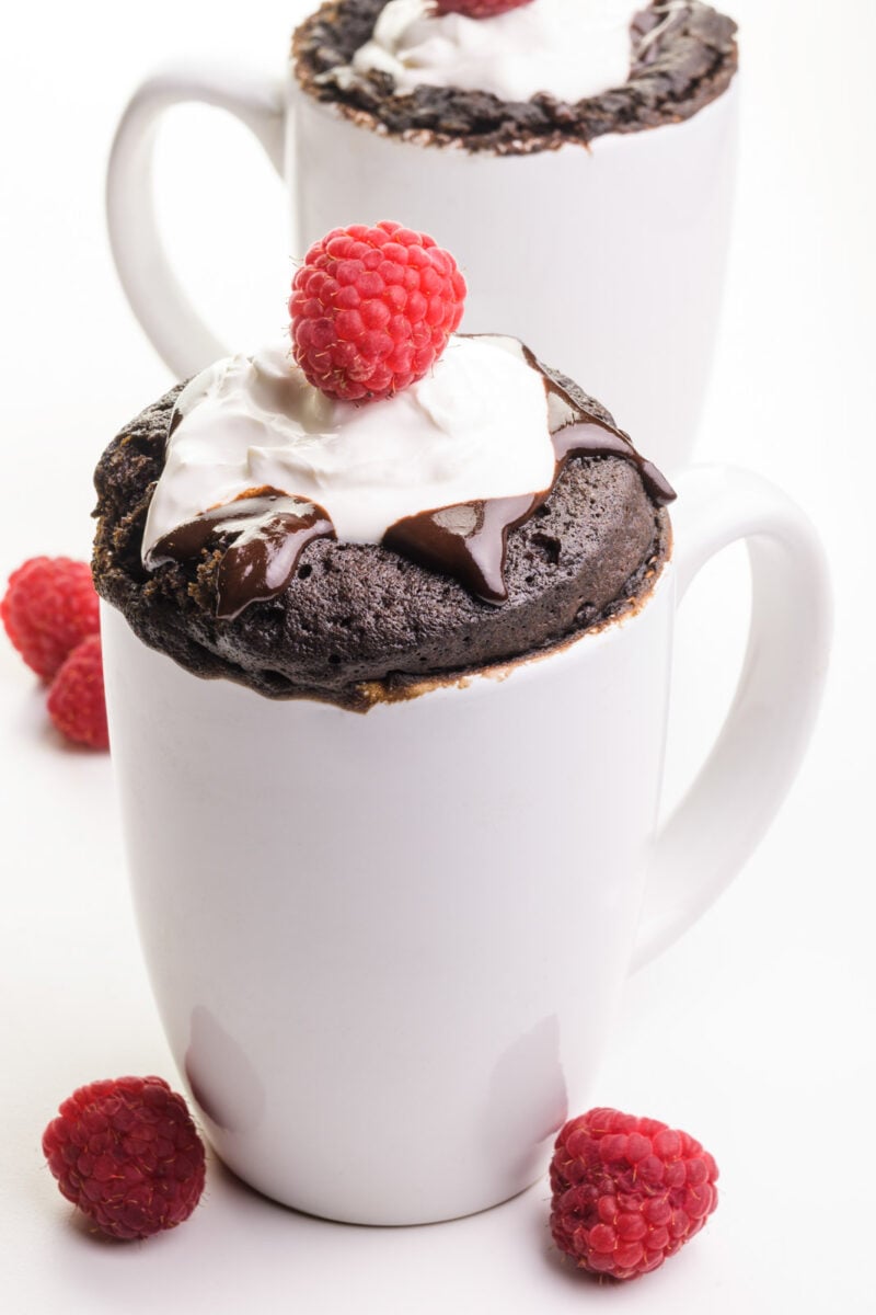 Two white coffee mugs hold chocolate vegan mug cakes with chocolate syrup, whipped cream, and raspberries on top. There are fresh raspberries on the table next to the mugs.