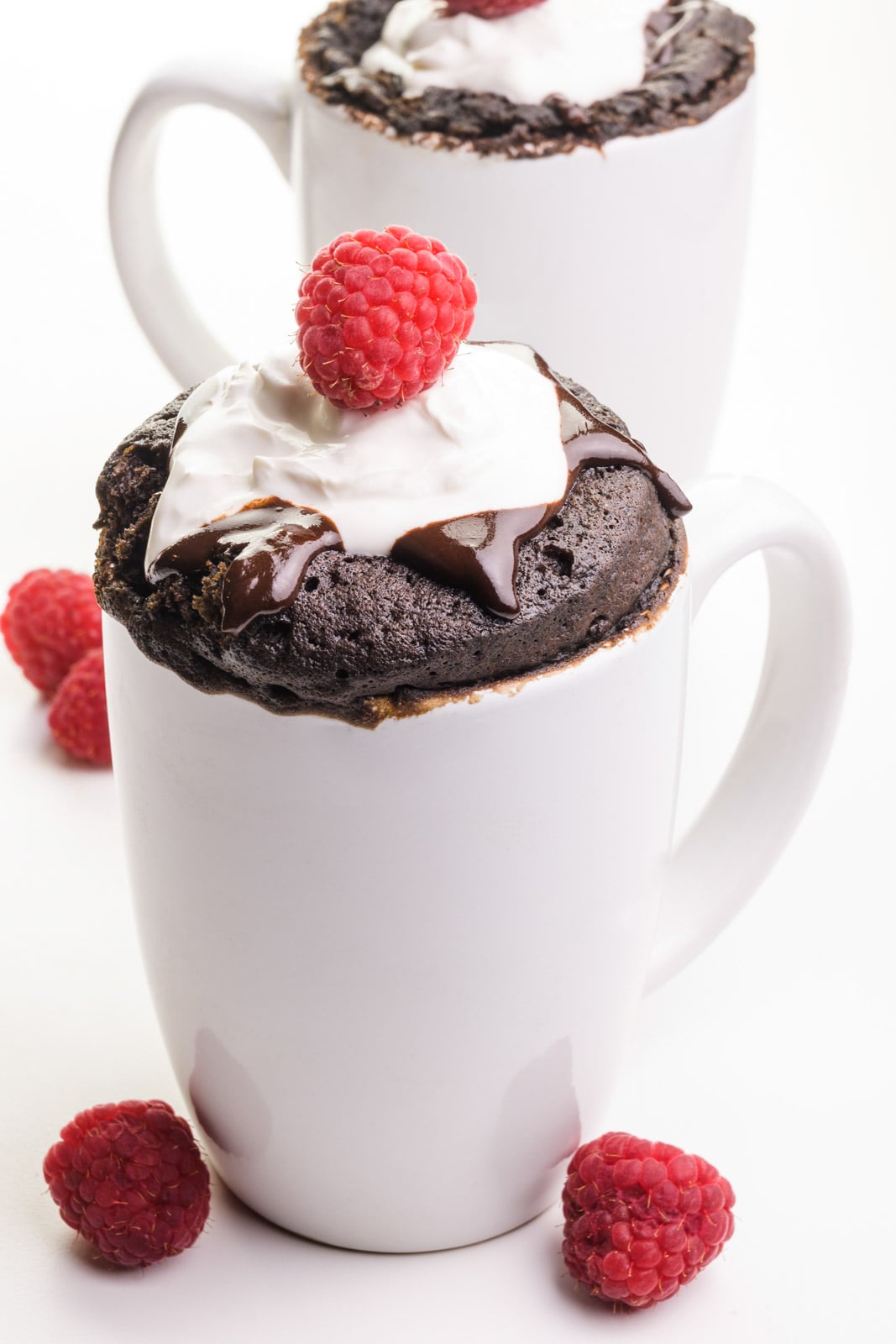 Two white coffee mugs hold chocolate vegan mug cakes with chocolate syrup, whipped cream, and raspberries on top. There are fresh raspberries on the table next to the mugs.