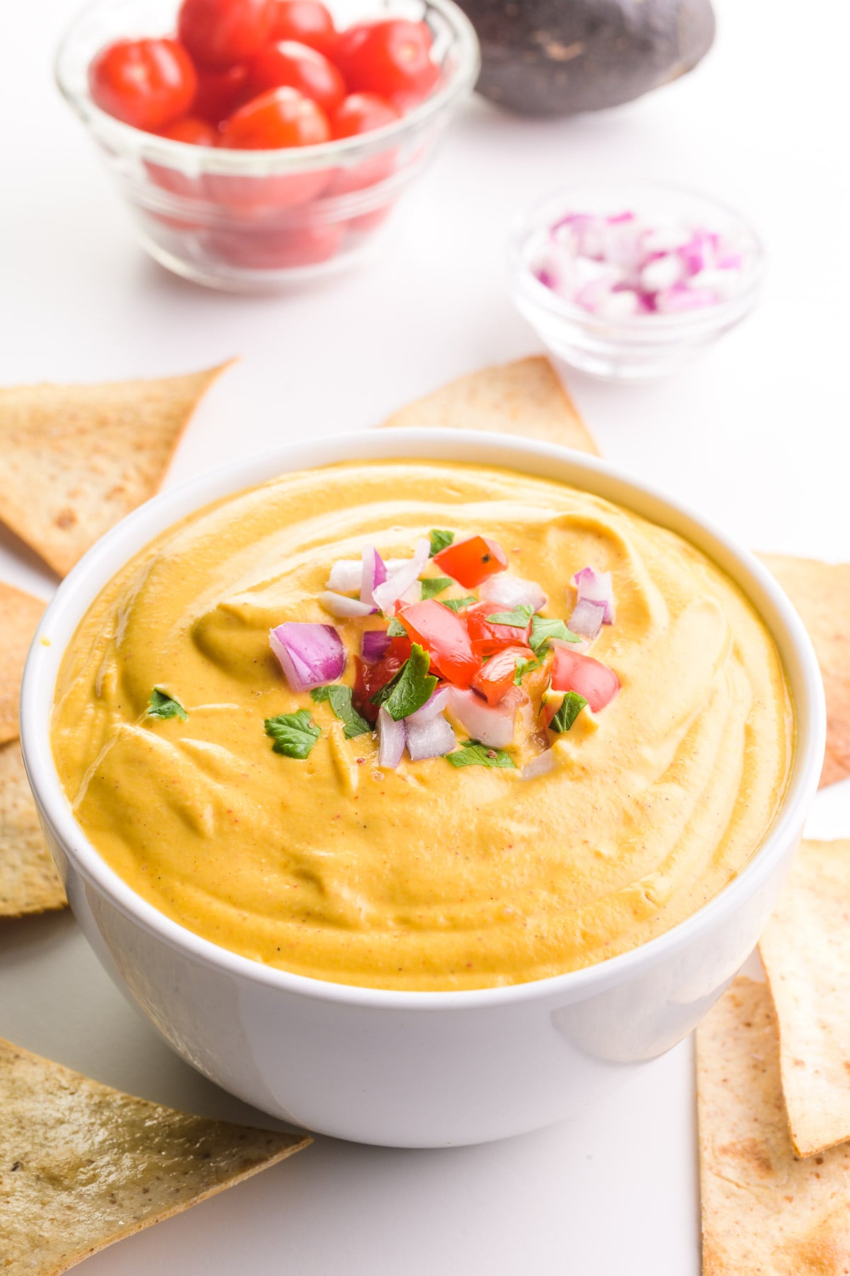 A bowl of golden sauce has chopped veggies on top. There are tortilla chips and more veggies in bowls around the sauce.